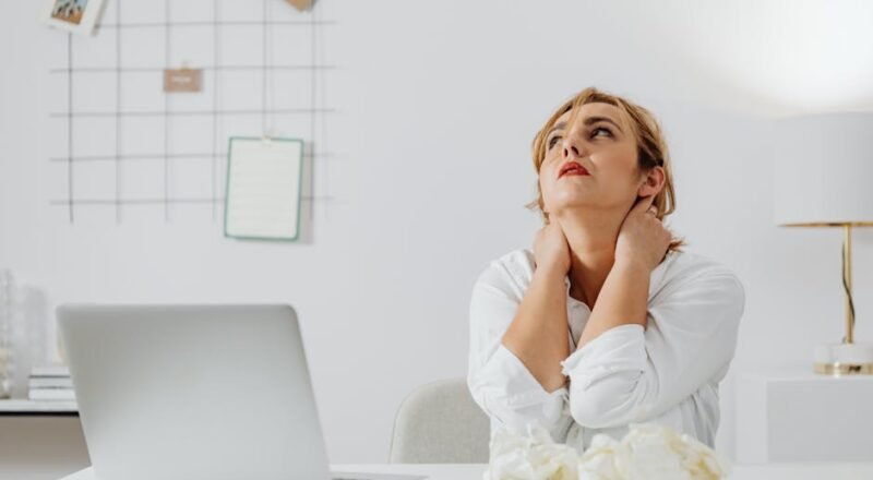 A stressed woman in white long sleeves sits at a desk with a laptop, looking upwards in an office.