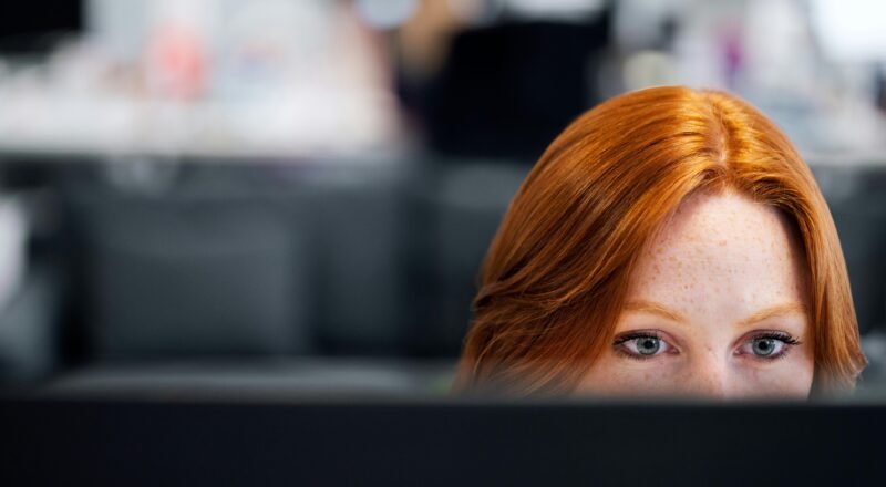 A woman with red hair intensely focused on a computer screen in an office setting.