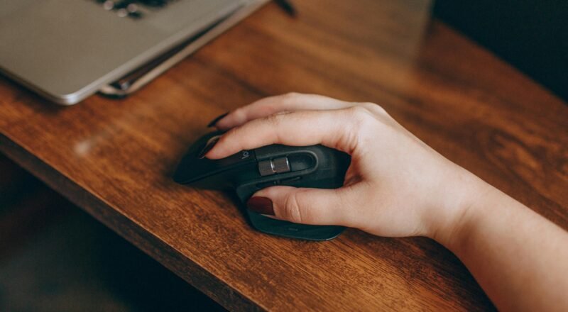 A close-up image of a hand with manicured nails using a wireless mouse on a wooden desk beside a laptop.