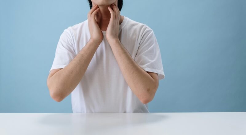 Close-up of an individual in a white crew neck t-shirt placing hands near neck, isolated on a blue backdrop.